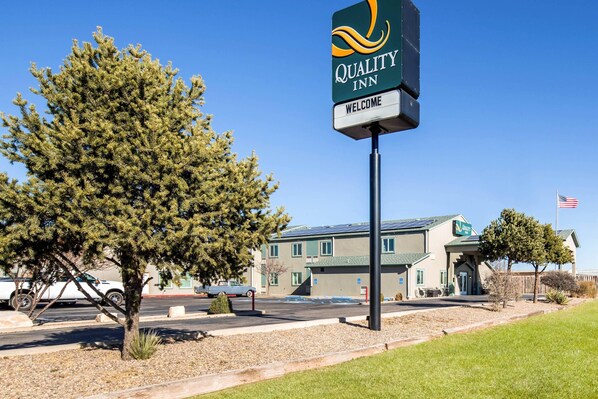 A Quality Inn sign with a welcoming message, a building with a green roof, and a parking lot with vehicles. - Quality Inn Moriarty (Moriarty)