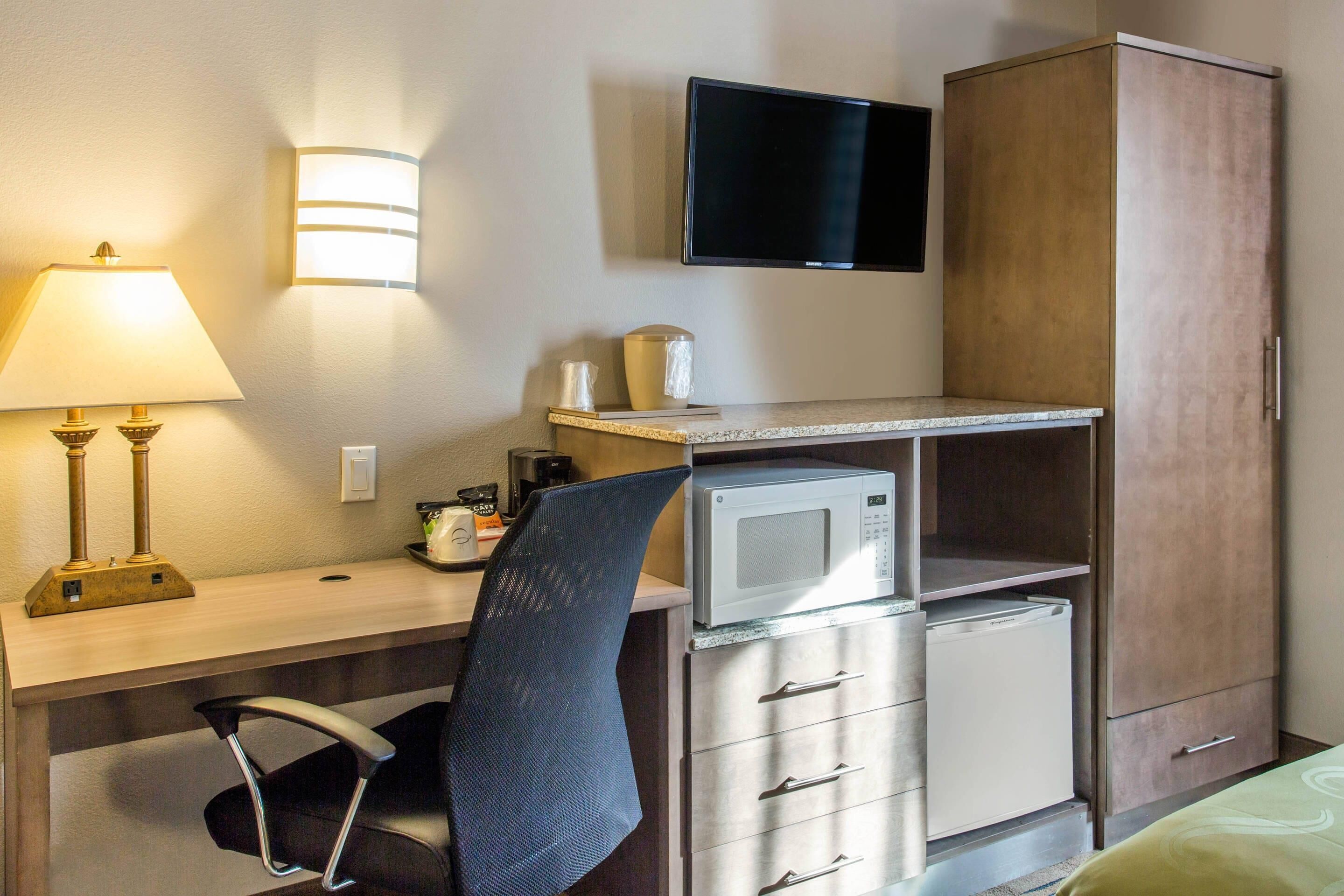 Quarto de hotel com escrivaninha, cadeira, micro-ondas, televisão e guarda-roupa.