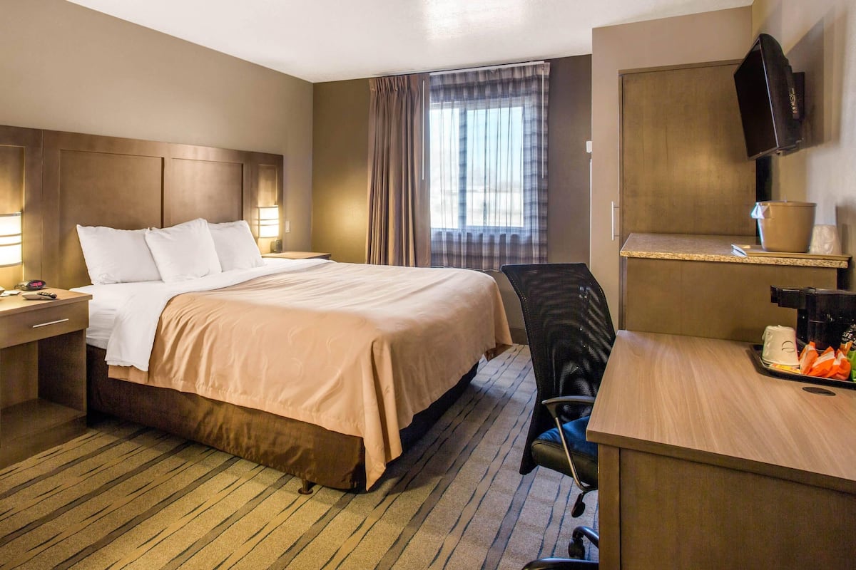 A hotel room with a large bed, a desk with a coffee maker, a TV mounted on the wall, and a window with curtains.