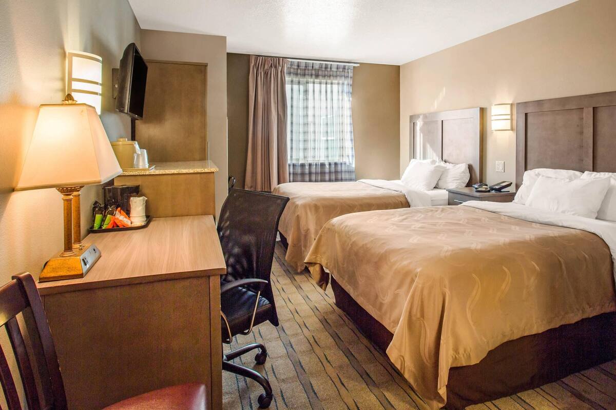 A hotel room with two beds, a desk with a lamp, a chair, a TV, and a window with curtains.