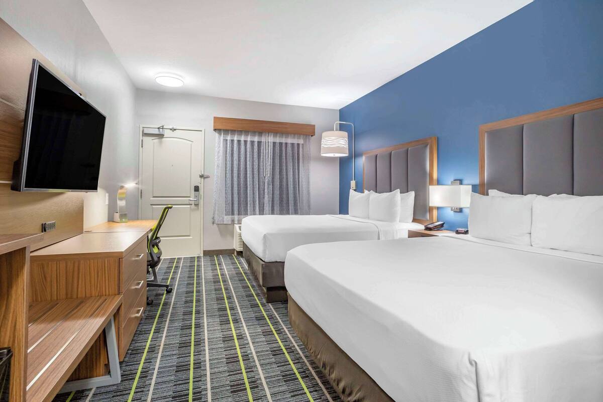 Quarto de hotel com duas camas, uma escrivaninha, televisão de tela plana, luminária de mesa e janela com cortinas.
