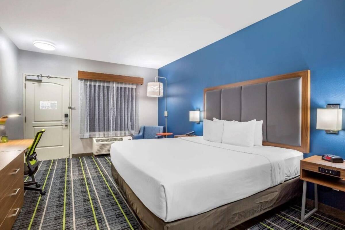 Quarto de hotel com uma cama grande, uma mesa com cadeira, uma janela com cortinas e uma luminária na parede.