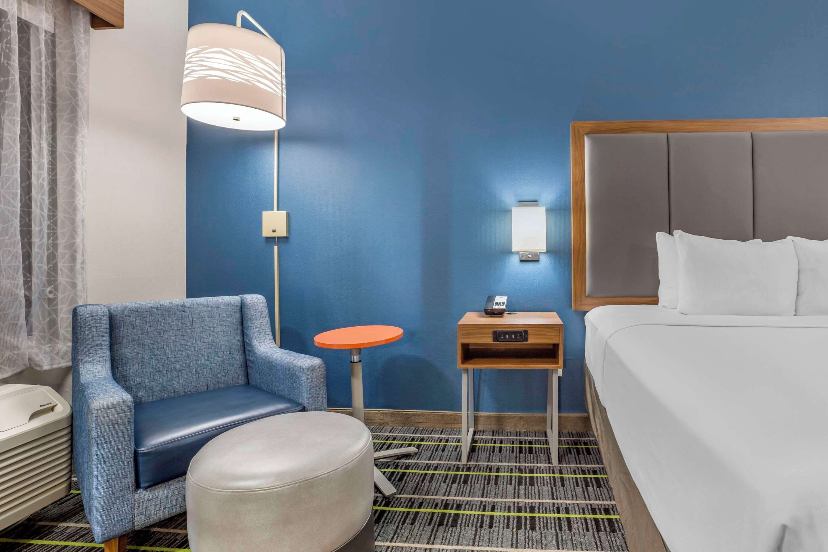 Habitació d'hotel amb una paret blava, un llit amb llençols blancs, un sofà blau, una taula rodona petita, una làmpada i una meseta de nit amb un telèfon.