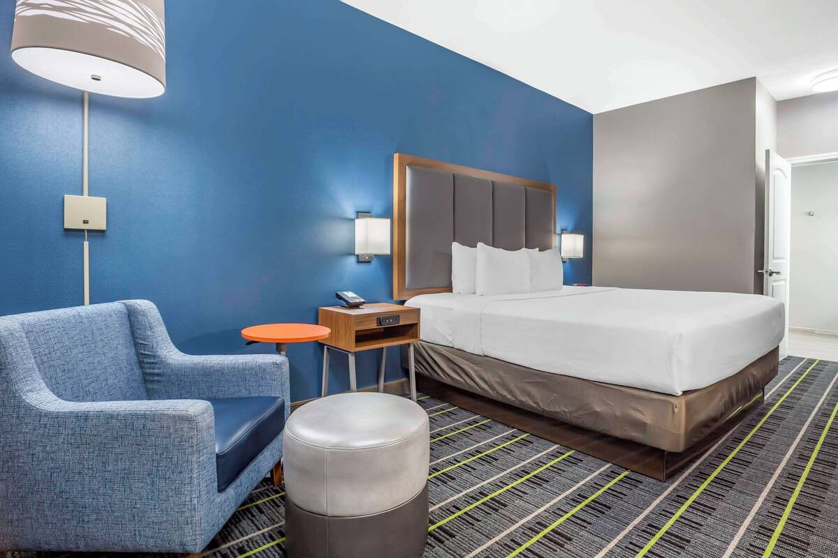 Quarto de hotel com cama, uma cadeira, mesinha, abajur e lixeira.