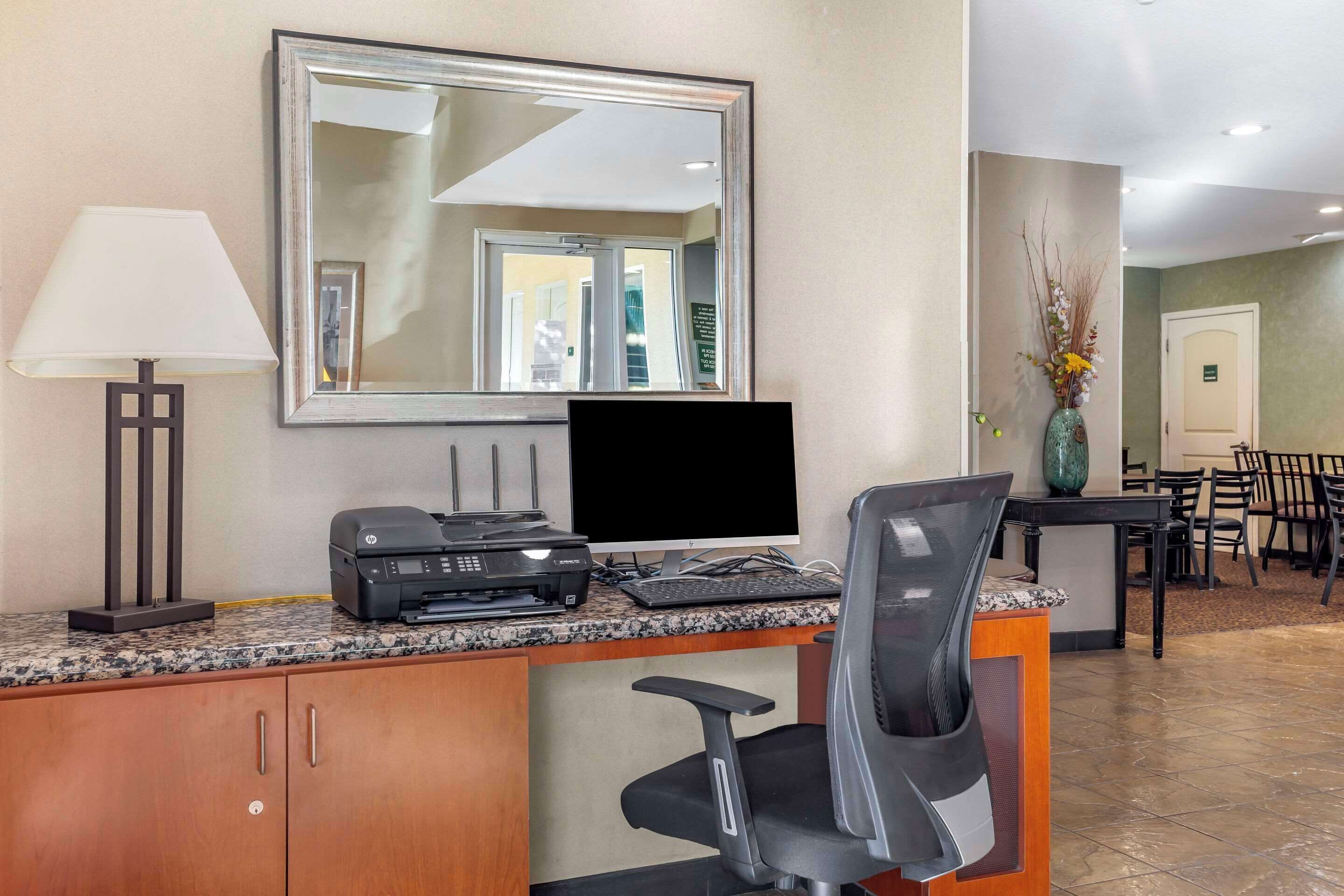 A hotel reception desk with a computer, printer, and a large mirror. There is a lamp, a vase with flowers, and a dining area in the background.