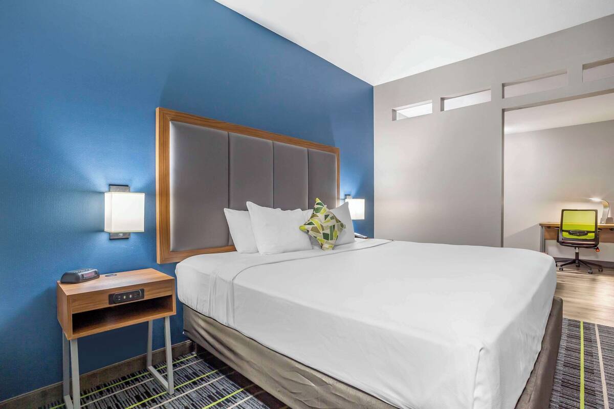 Quarto de hotel com uma cama grande, uma mesa de cabeceira de madeira, uma escrivaninha com cadeira e uma luminária de mesa.