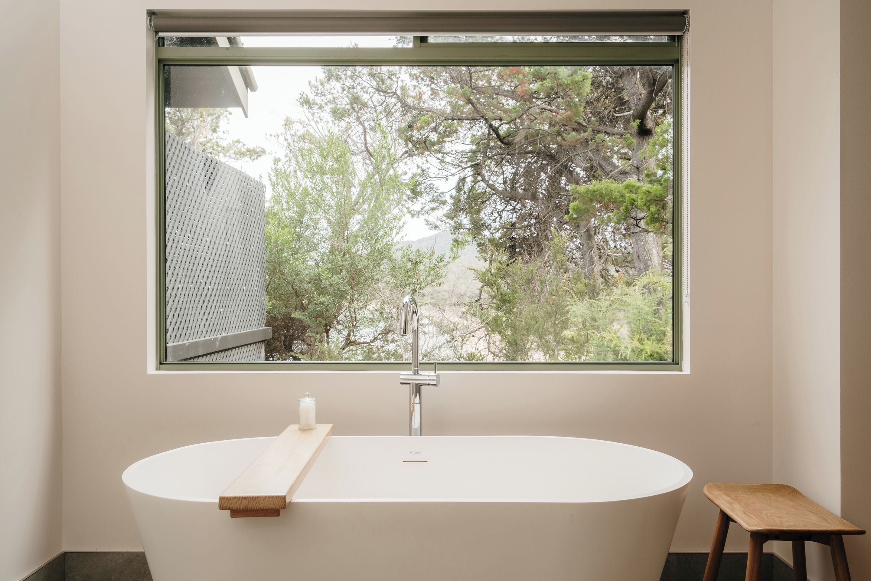 Un baño moderno con una bañera independiente, un taburete de madera y una ventana con vistas a árboles.