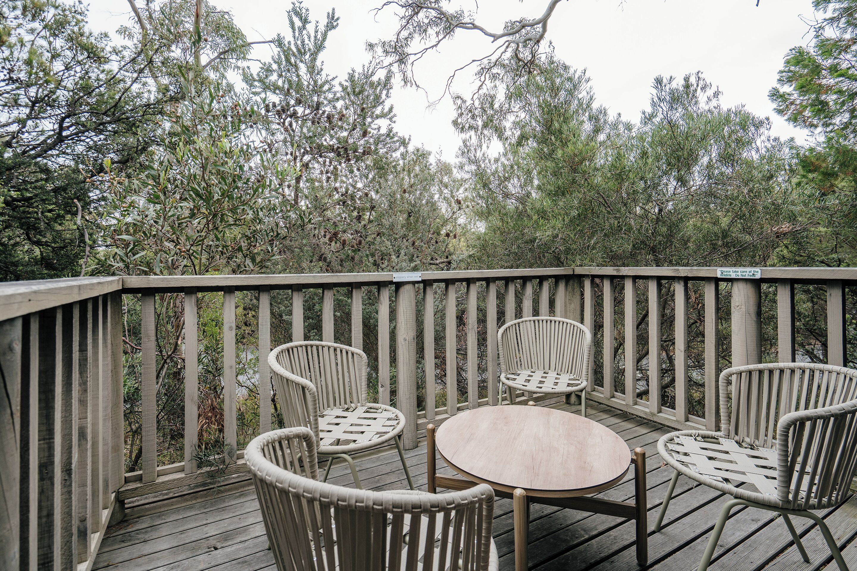 A wooden deck with wicker chairs and a round table, surrounded by trees.