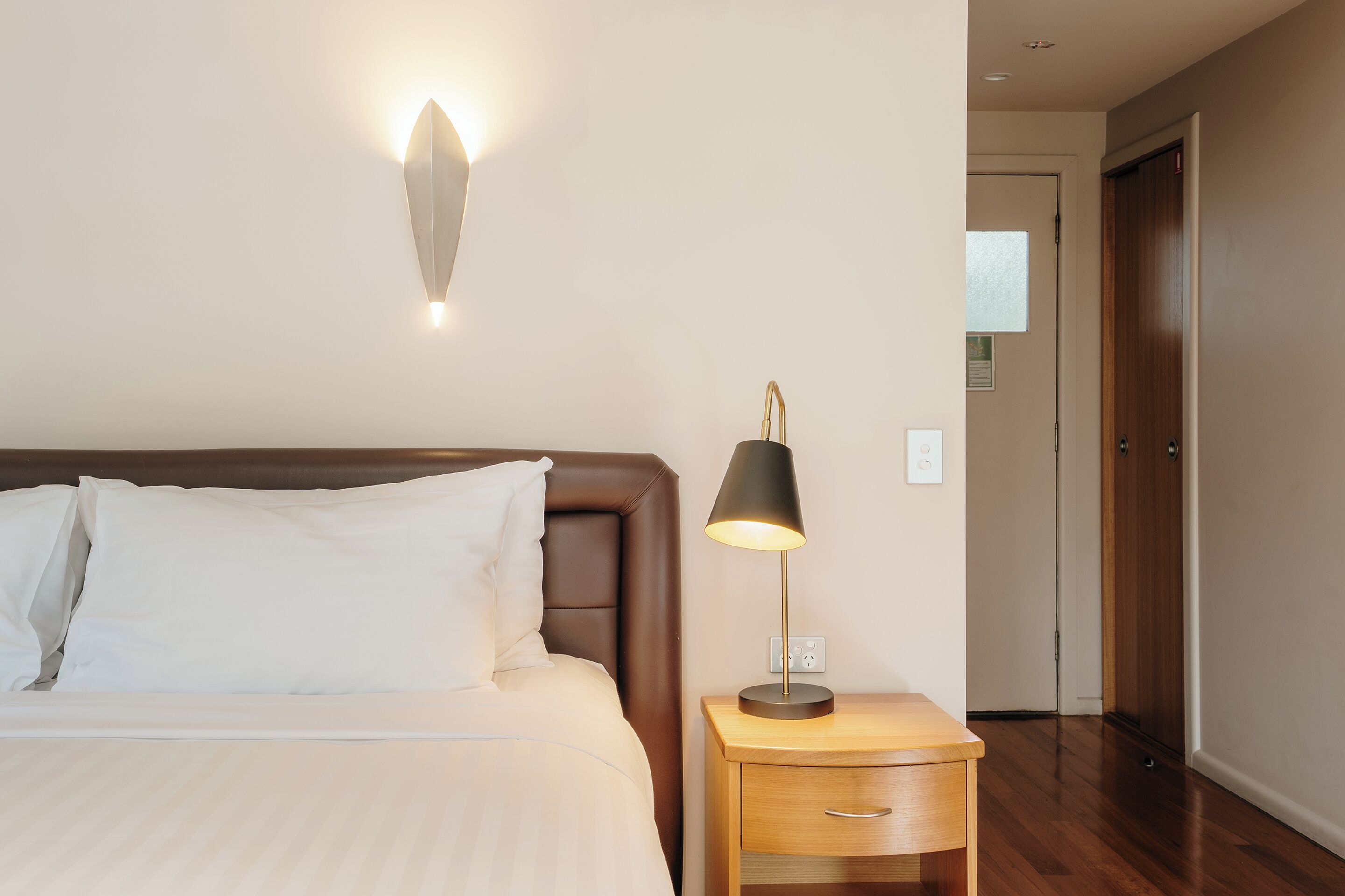 Una cama bien hecha con sábanas blancas, una lámpara de noche y una lámpara de pared.