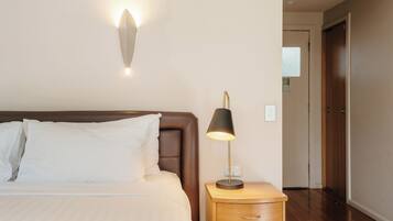Una cama bien hecha con sábanas blancas, una lámpara de noche y una lámpara de pared.