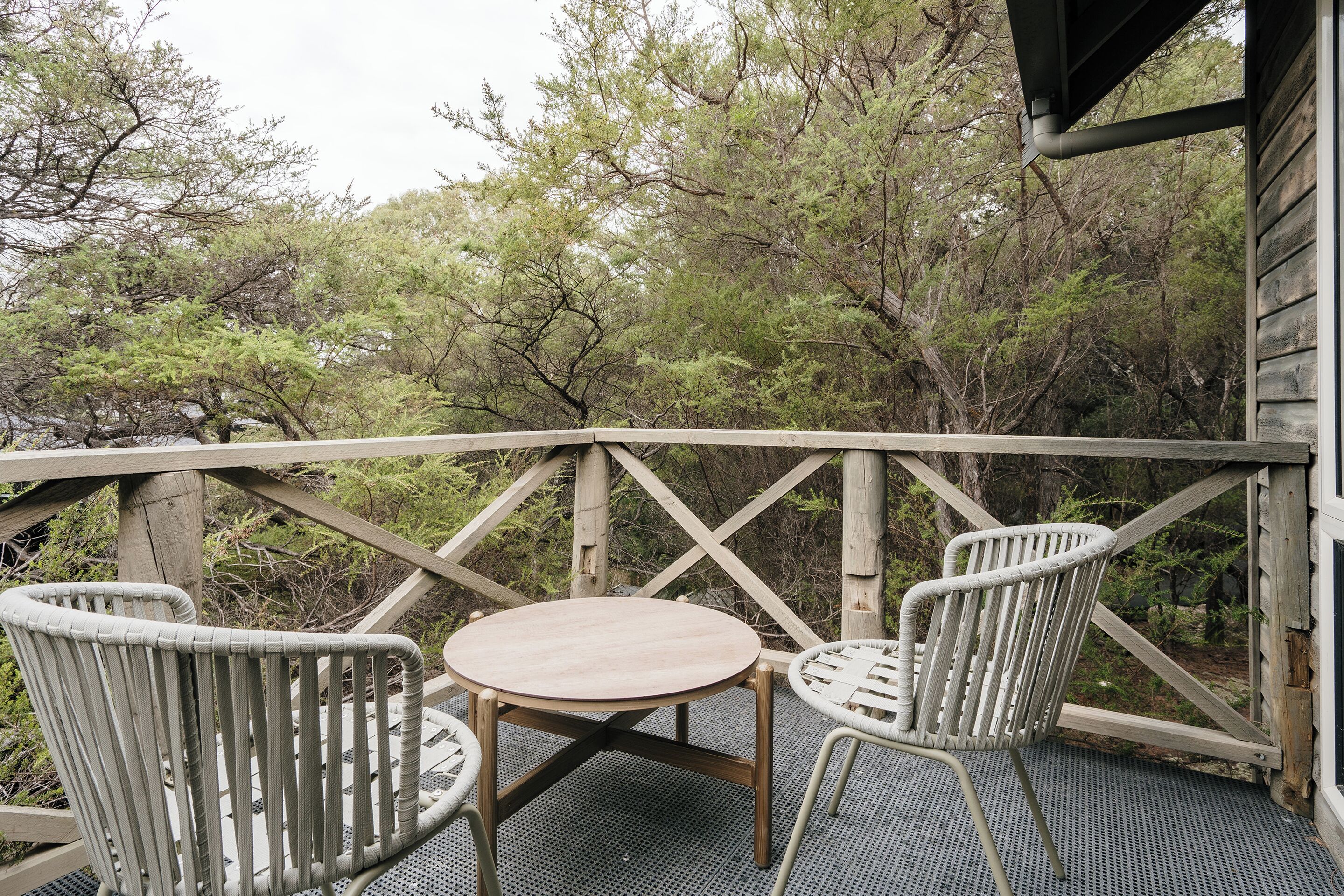 A wooden deck with a round table and two wicker chairs, surrounded by trees.
