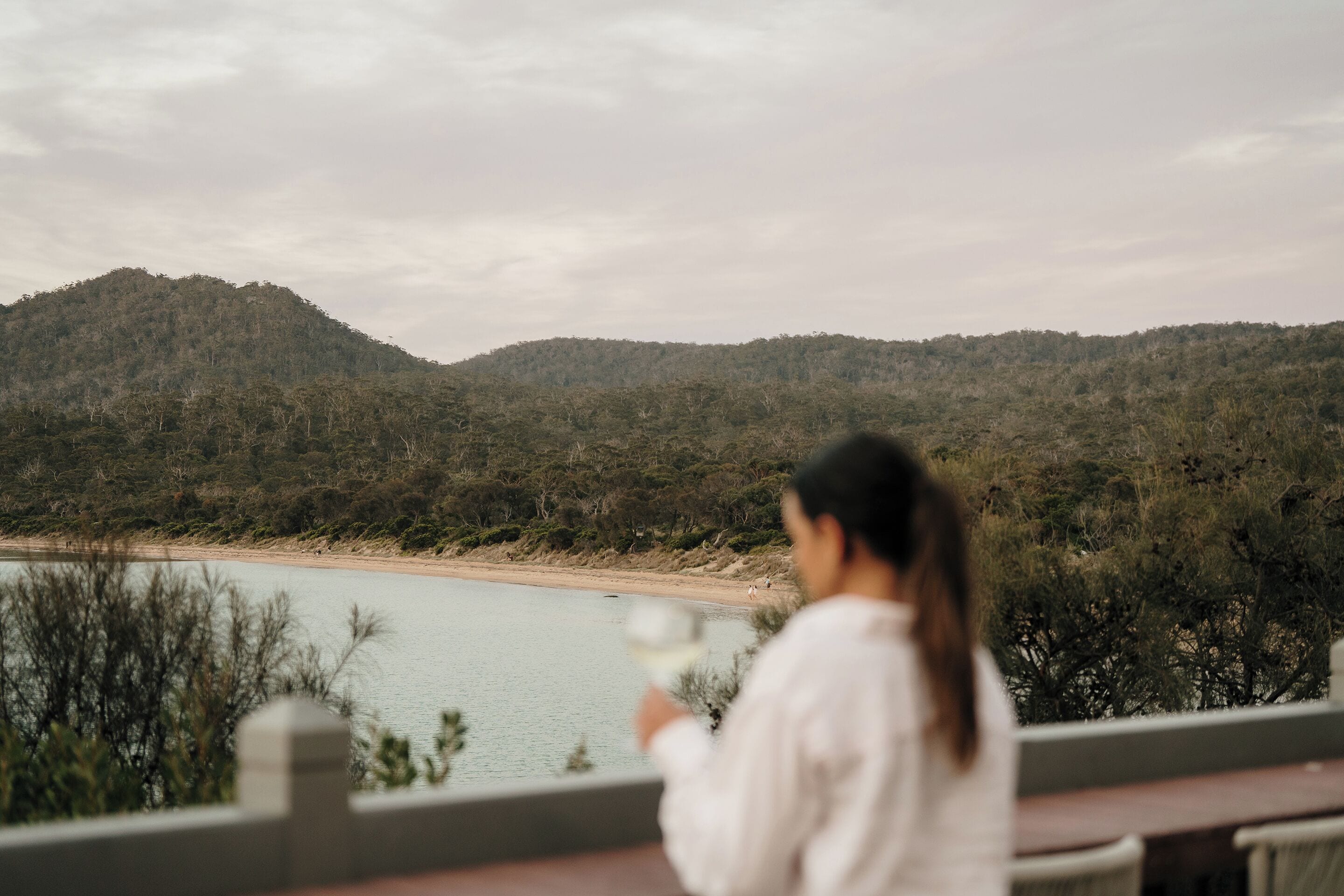 A woman holding a glass, overlooking a lake with a forested hill in the background.