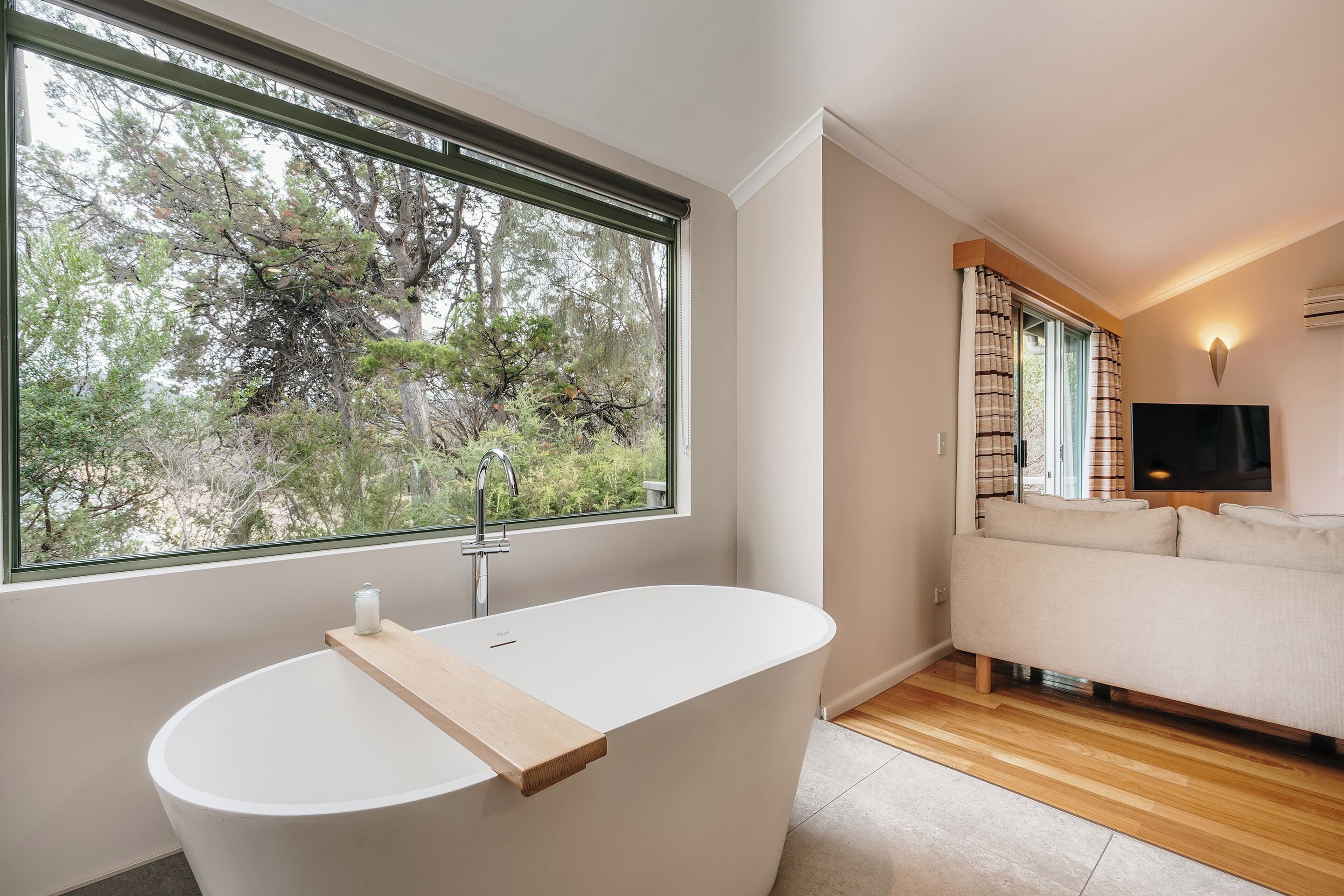 Une salle de bain moderne avec une baignoire sur pieds, une grande fenêtre donnant sur les arbres et un canapé dans la pièce adjacente.