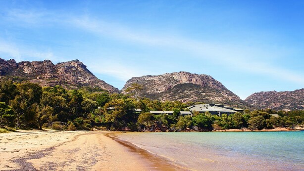 A beach with clear waters, a sandy shore, and a house surrounded by trees and mountains in the background. - Freycinet Lodge (Coles Bay)