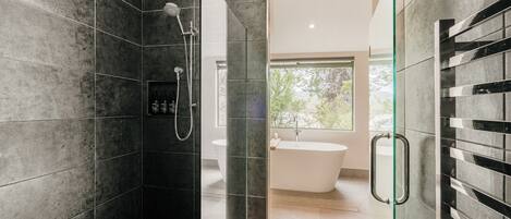 A modern bathroom with dark tiled walls, a glass shower enclosure, and a white bathtub.