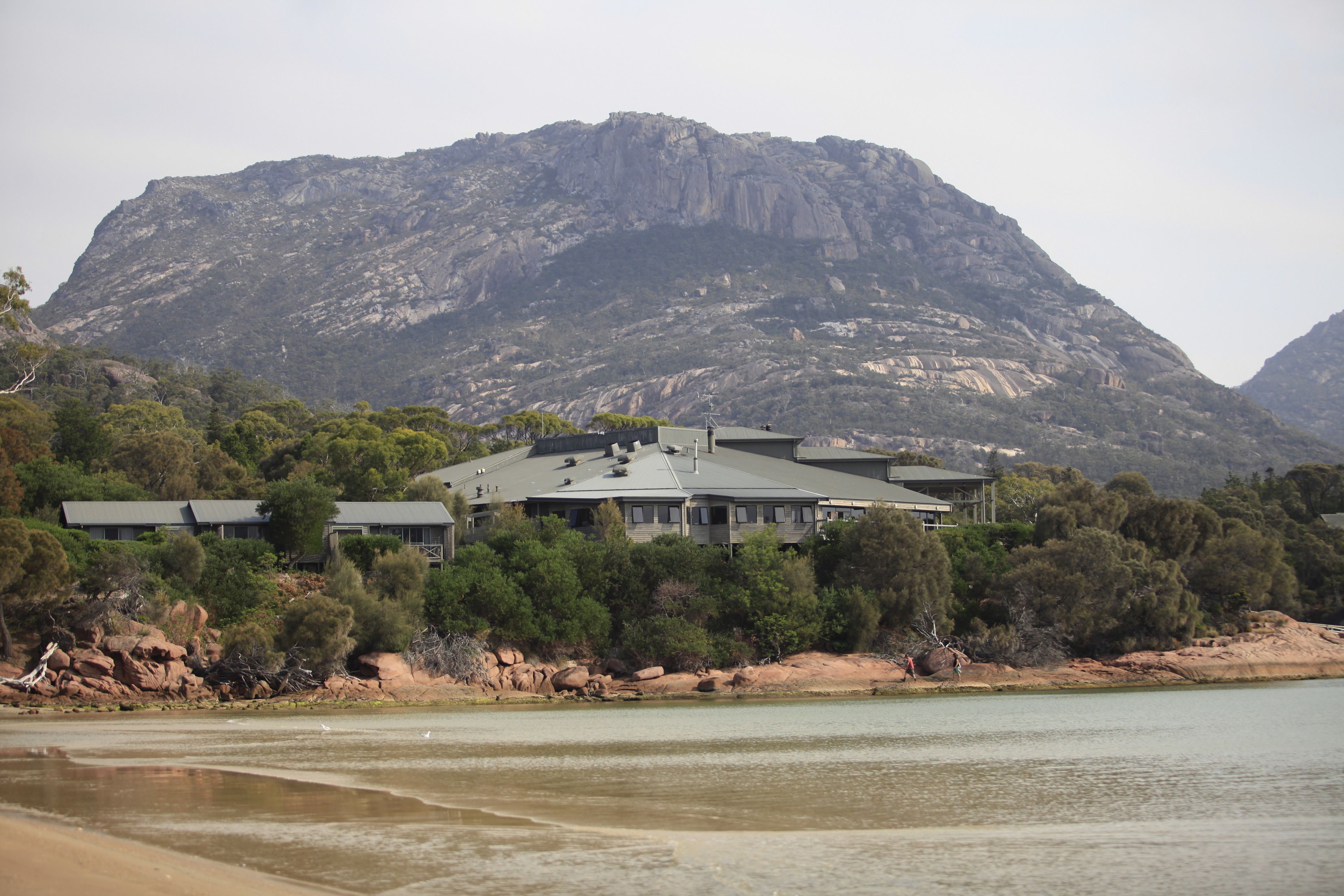 A large, modern building with a flat roof and multiple windows, situated near a body of water with a mountainous backdrop.