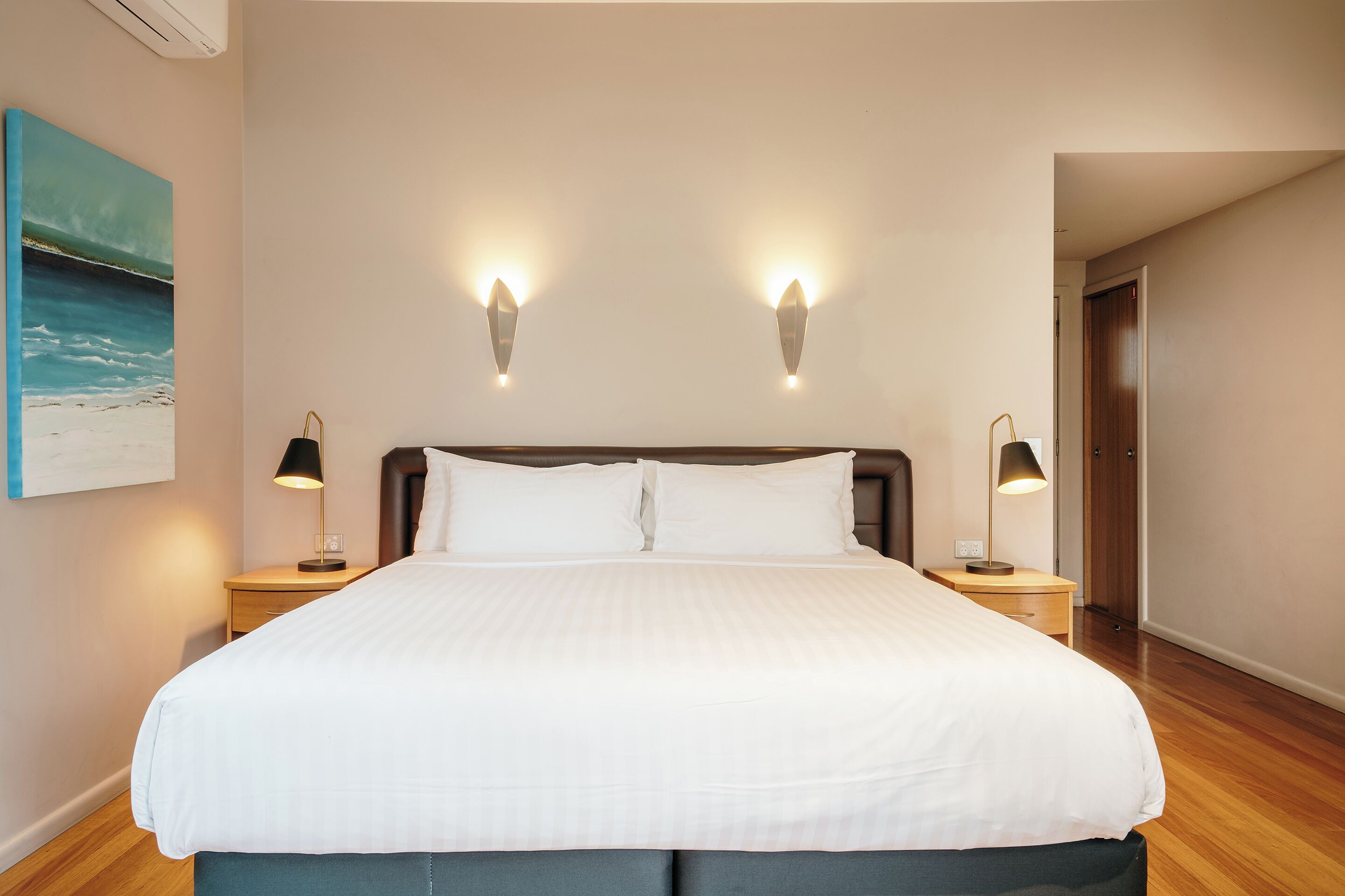 Ein ordentlich gemachtes Bett mit weißer Bettwäsche, zwei Nachttischlampen und ein Strandbild an der Wand.