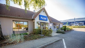 Een hotel genaamd 'Kyriad' met een modern design, een parkeerplaats en een Peugeot-autohandel op de achtergrond.