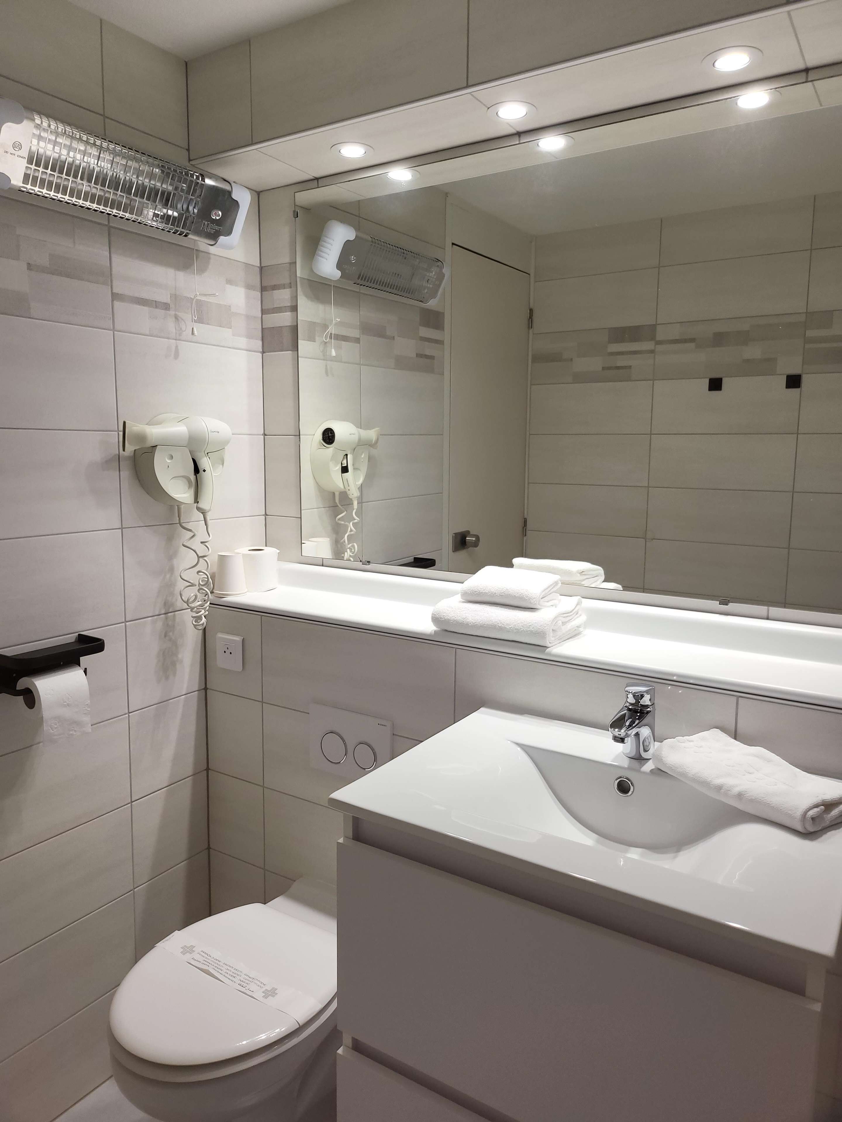 A modern bathroom with a white sink, mirror, and toilet. There are two hair dryers mounted on the wall, and a towel rack with folded towels.