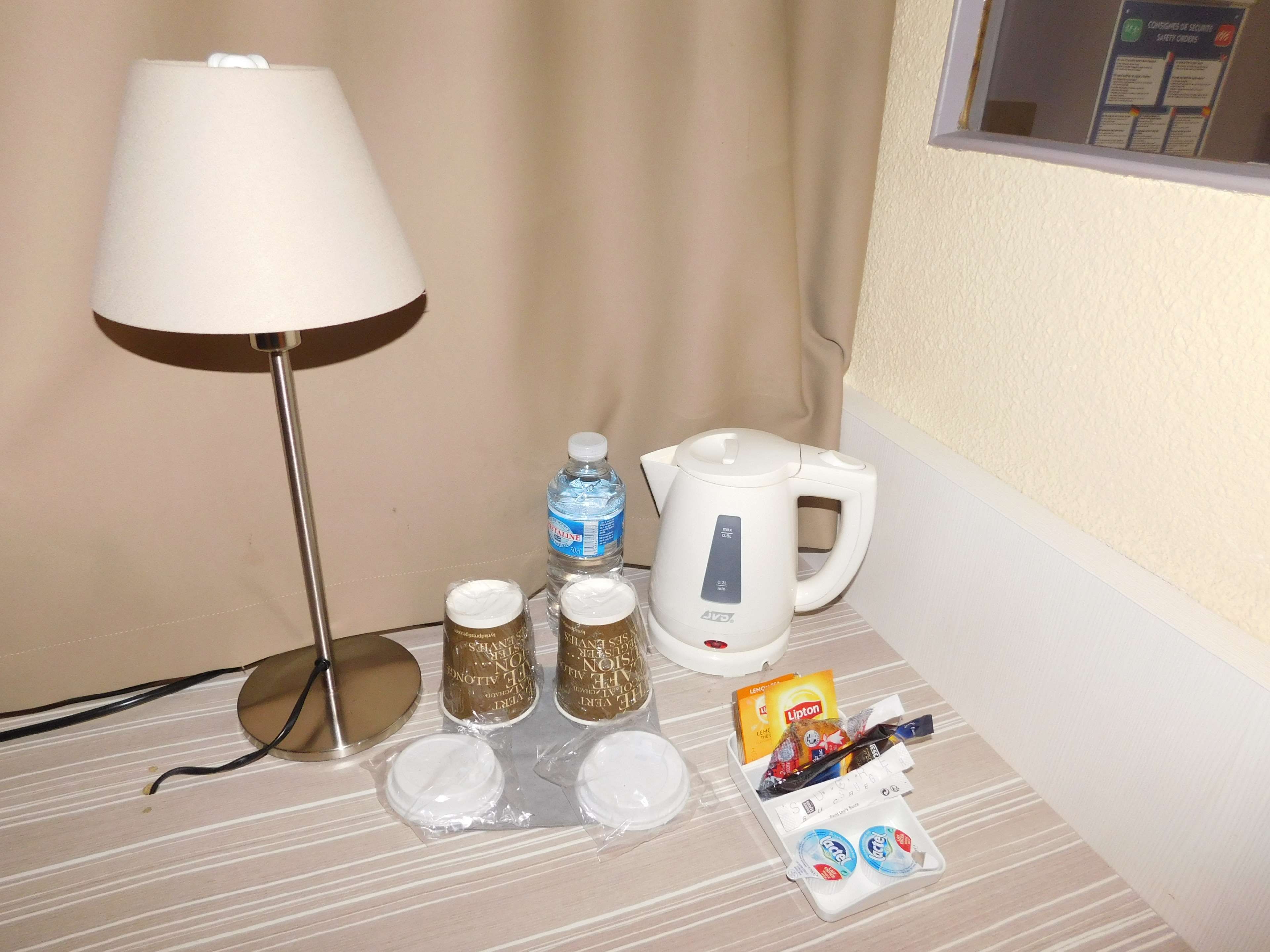 Una habitación de hotel con una lámpara, una tetera, una botella de agua y algunos artículos de aseo sobre la mesa.