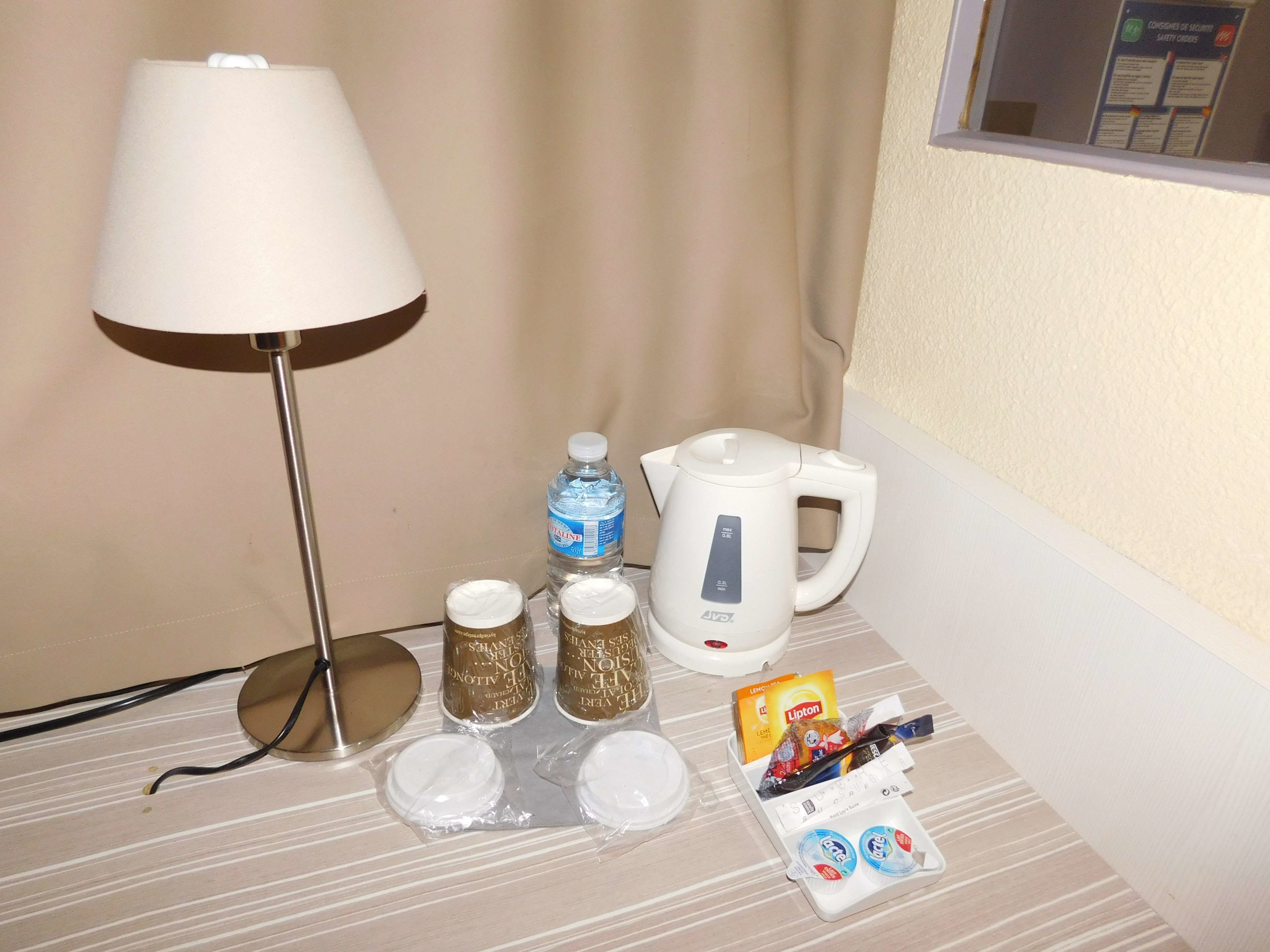 A hotel room with a lamp, a kettle, a bottle of water, and some toiletries on the table.