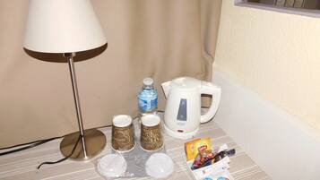 A hotel room with a lamp, a kettle, a bottle of water, and some toiletries on the table.