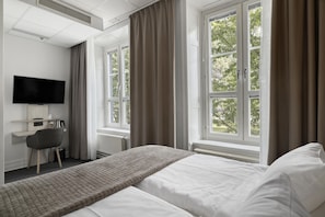 Habitación de hotel con cama, escritorio, silla, televisor y ventanas grandes con cortinas.