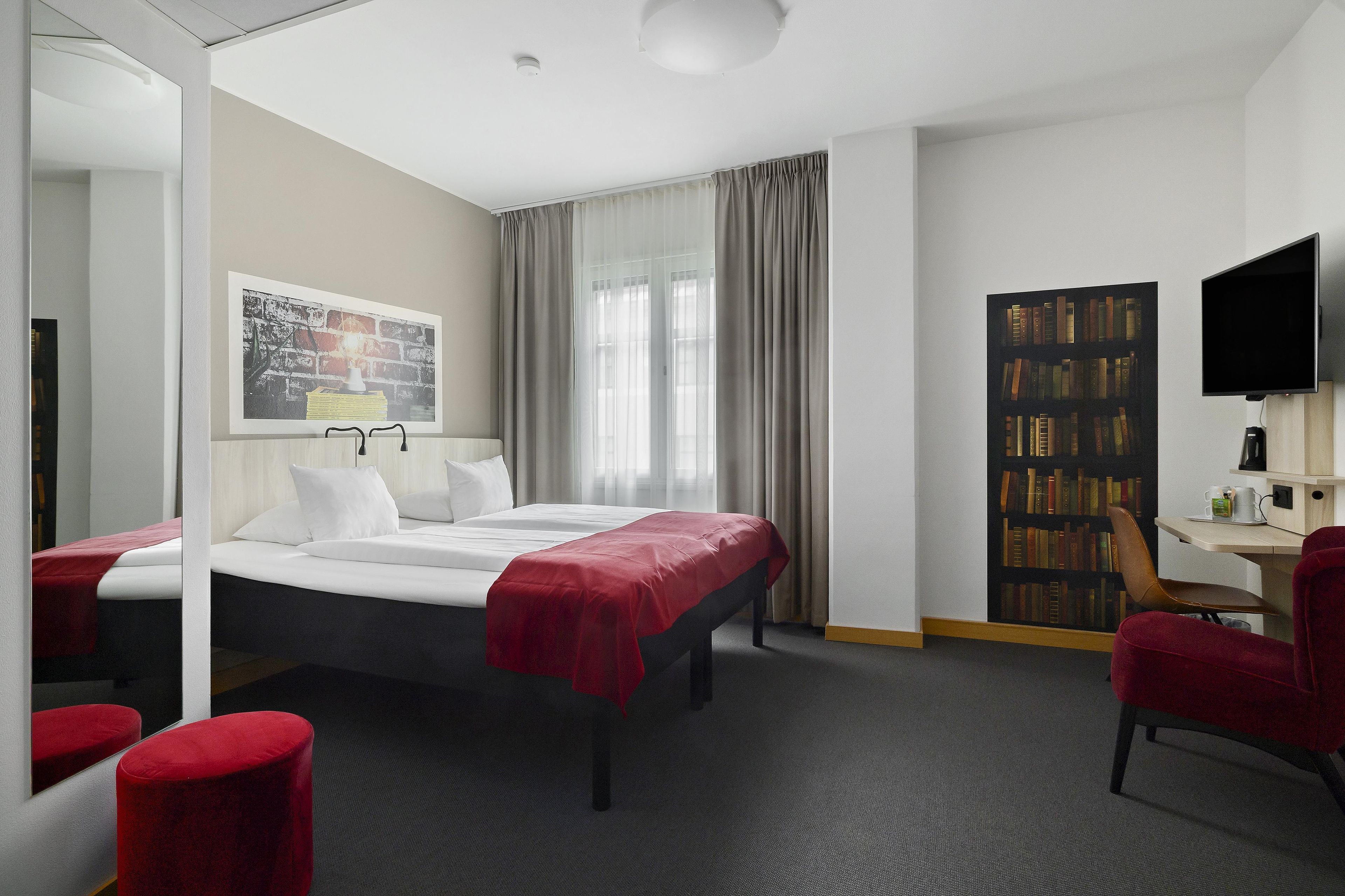 A hotel room with a bed, a desk, a chair, a bookshelf, and a television.