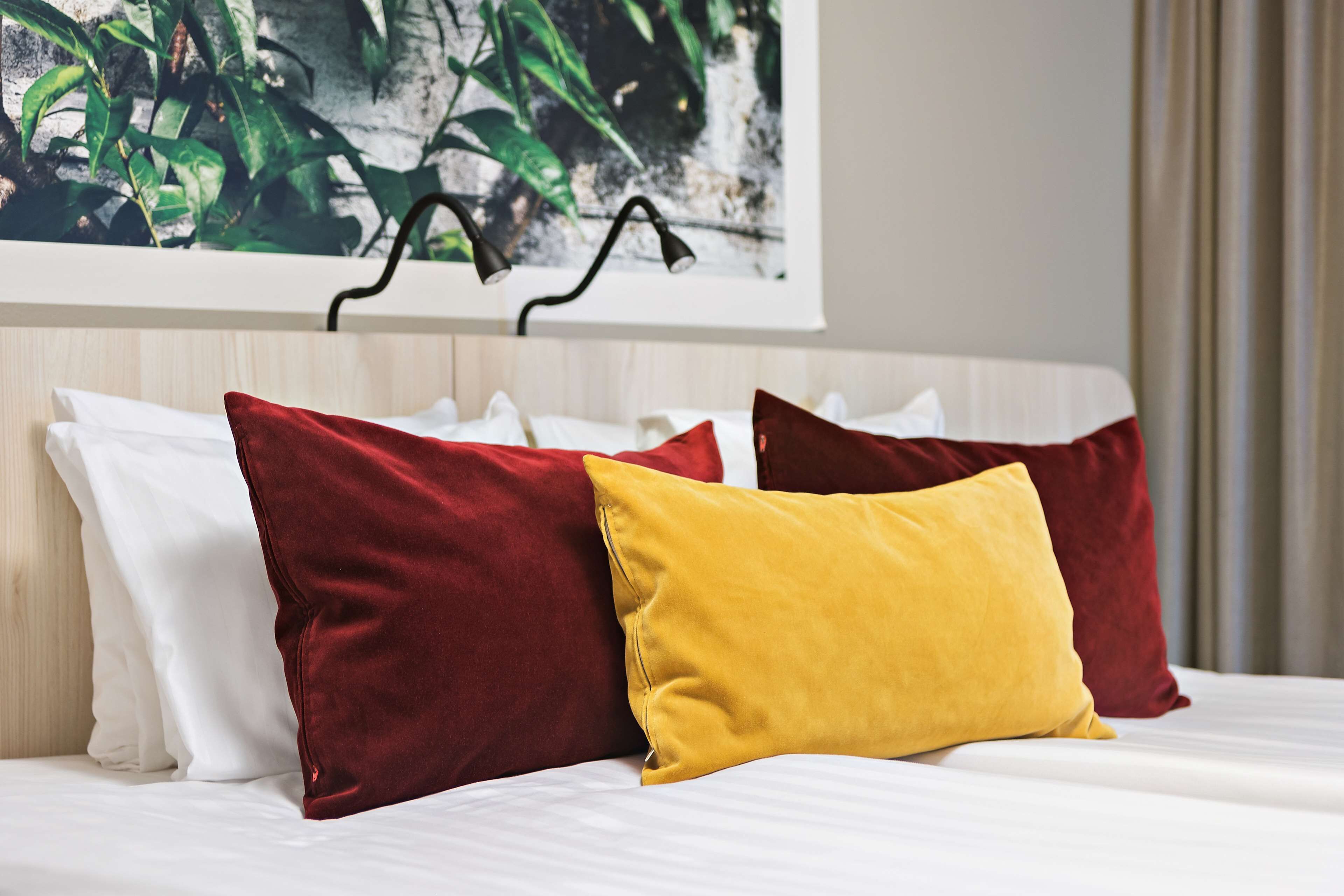 Una cama con almohadas blancas y rojas, una almohada amarilla y un cuadro con hojas verdes en la pared.