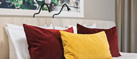 Una cama con almohadas blancas y rojas, una almohada amarilla y un cuadro con hojas verdes en la pared.