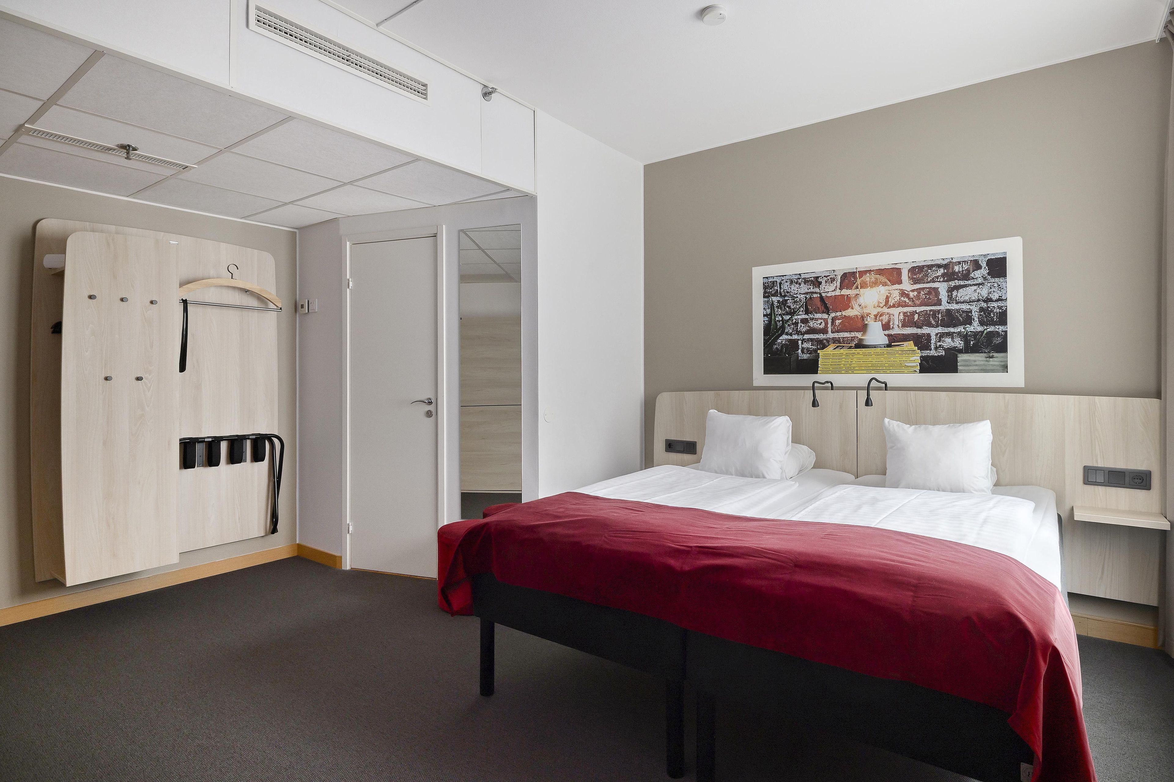 A hotel room with a bed, a wardrobe, a coat rack, and a wall-mounted light fixture.