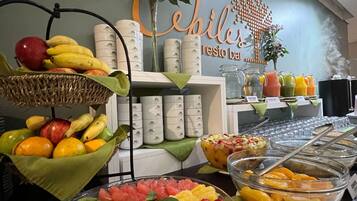 Un bufé con diversas frutas y bebidas, una pared con un letrero que dice "Los Chiles resto bar", y una planta decorativa.