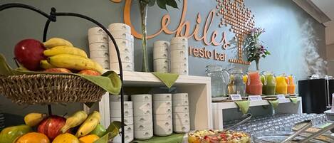 A buffet with various fruits and beverages, a wall with a sign reading "Los Chiles resto bar", and a decorative plant.