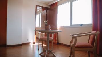 Una habitación con piso de madera, una mesa con un florero, una silla y una ventana con cortinas.