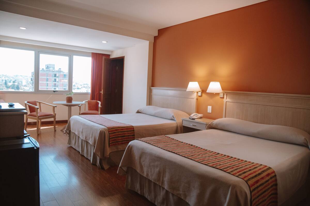 A hotel room with two beds, a desk, and a chair. There is a window with a view of buildings outside.