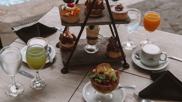 A poolside table with a variety of desserts and drinks, including a three-tiered dessert stand, a bowl of fruit, and several glasses of beverages. - Casa Real (Salta)