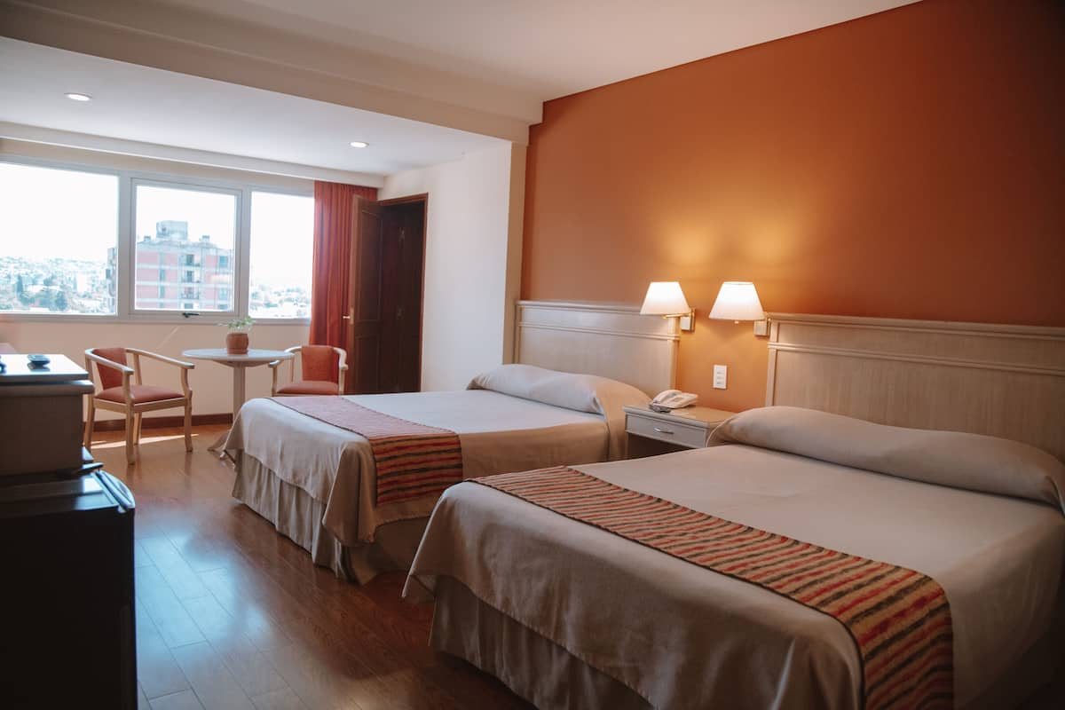 A hotel room with two beds, a desk, a chair, and a window with a city view.