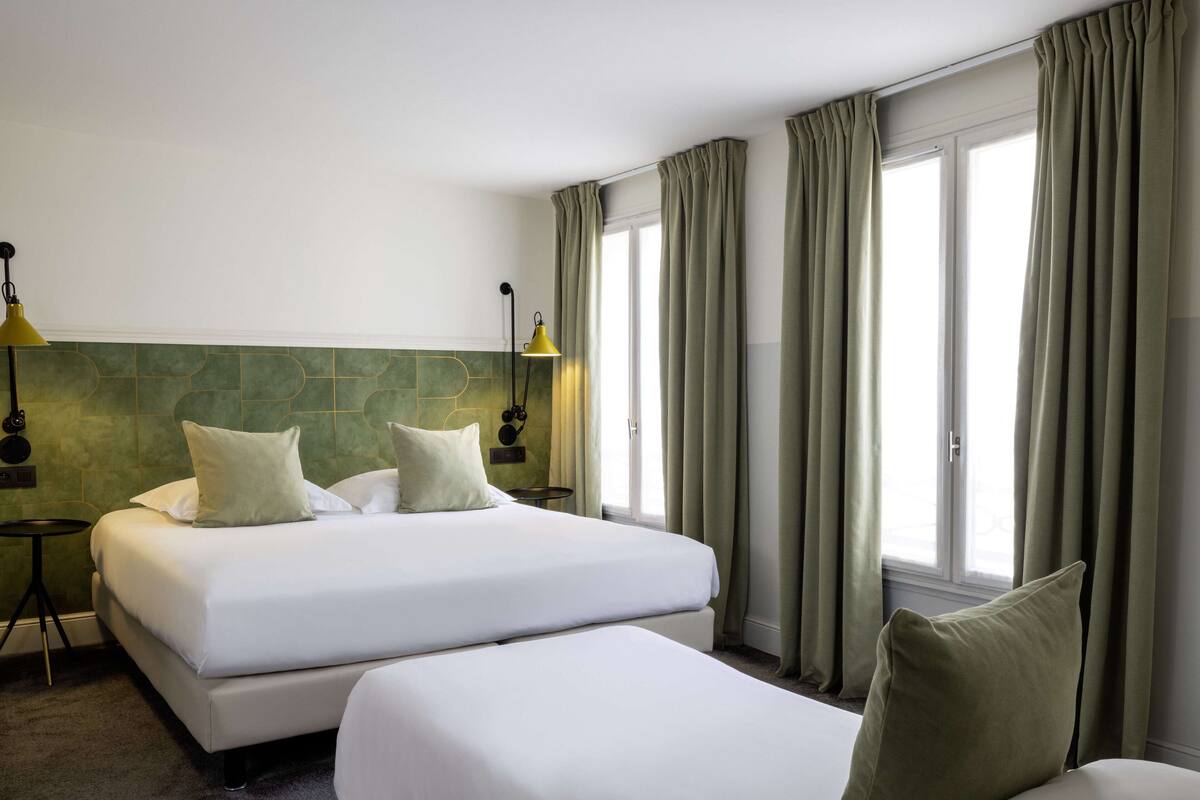 A hotel room with two beds, a green tiled headboard, a small table, and a lamp.