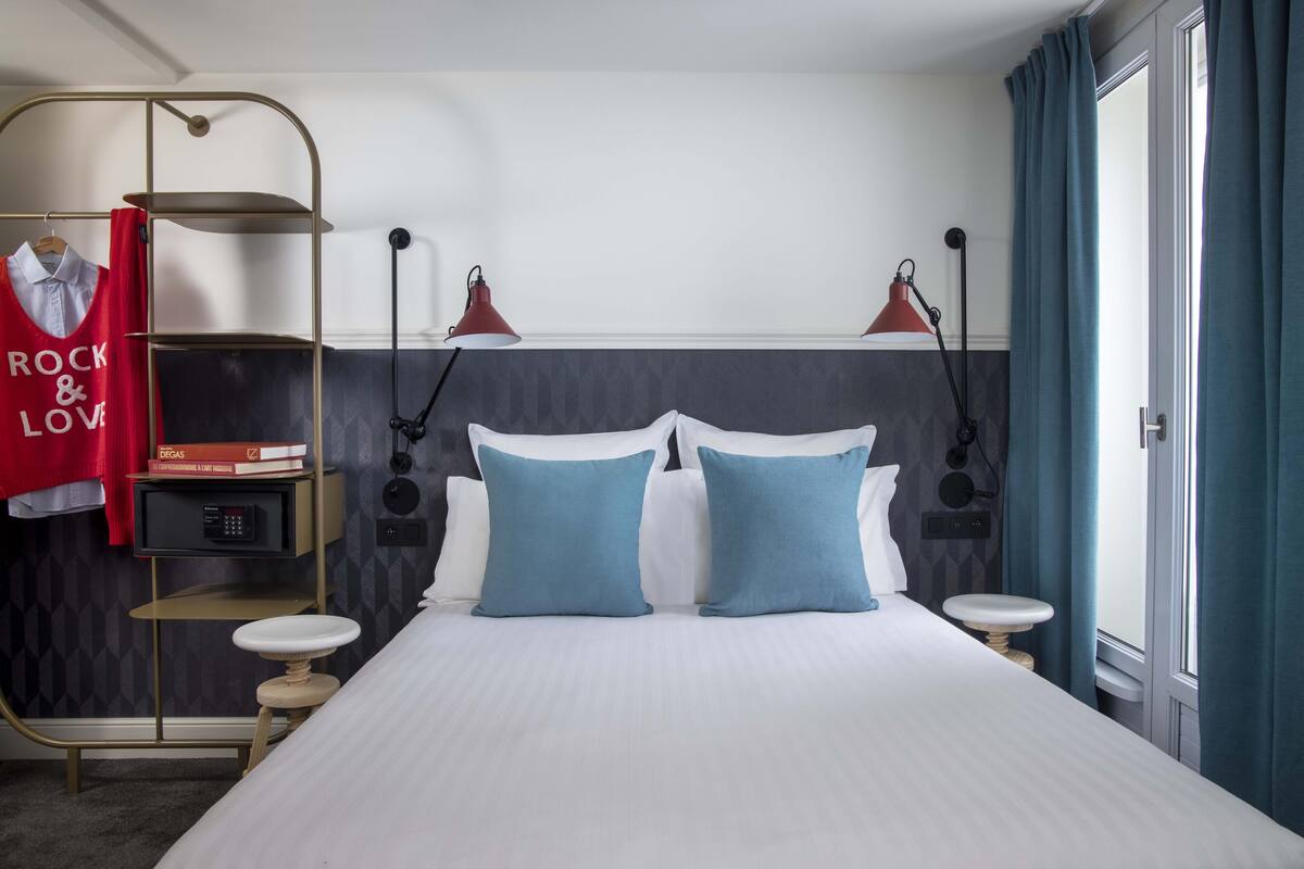 A neatly arranged hotel room with a bed, bedside tables, a shelf with books, a hanging garment, and a window with curtains.
