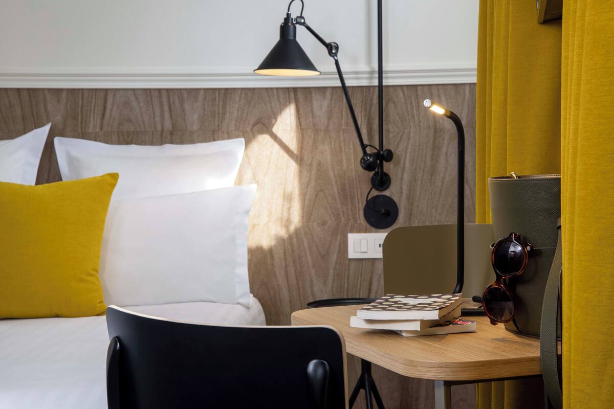 A neatly arranged hotel room with a bed, a desk, a chair, a lamp, and a pair of sunglasses on the wall.