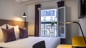 A hotel room with a bed, a desk, a chair, a lamp, and a window with a view of buildings and a balcony.