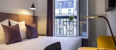 A hotel room with a bed, a desk, a chair, a lamp, and a window with a view of buildings and a balcony.