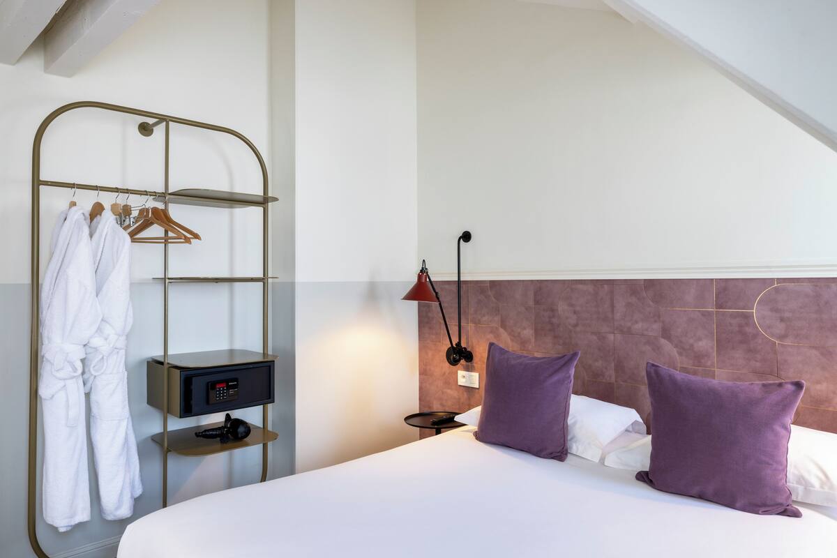 A hotel room with a bed, a nightstand, a robe rack, and a wall-mounted lamp.