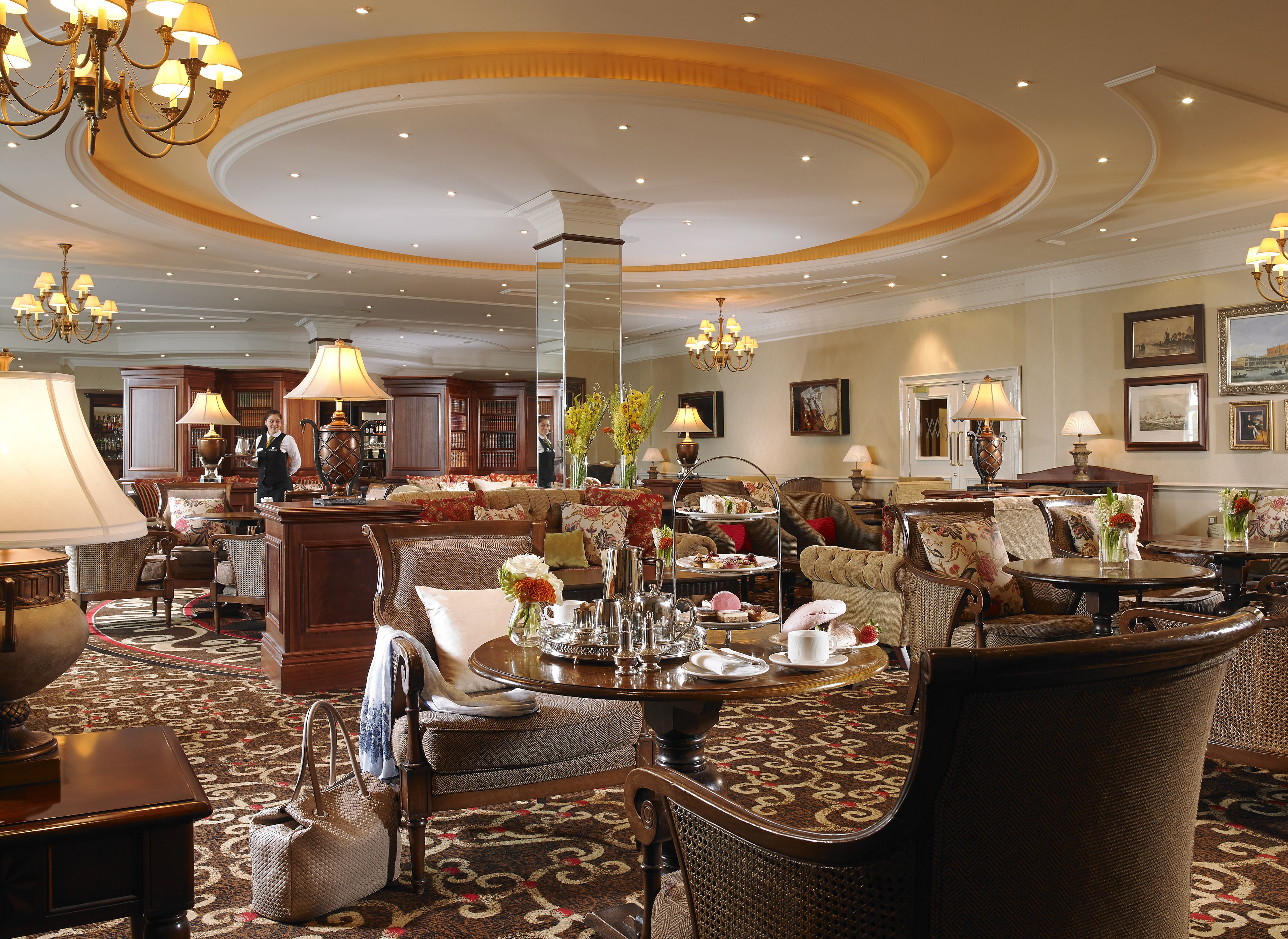 A spacious hotel lobby with a central seating area, multiple chandeliers, and a variety of seating options including sofas and armchairs.