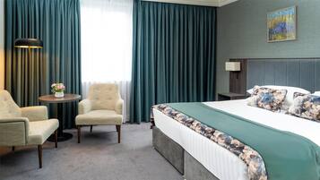 Habitación de hotel con una cama grande, una silla, una mesita con un jarrón de flores y un cuadro en la pared.