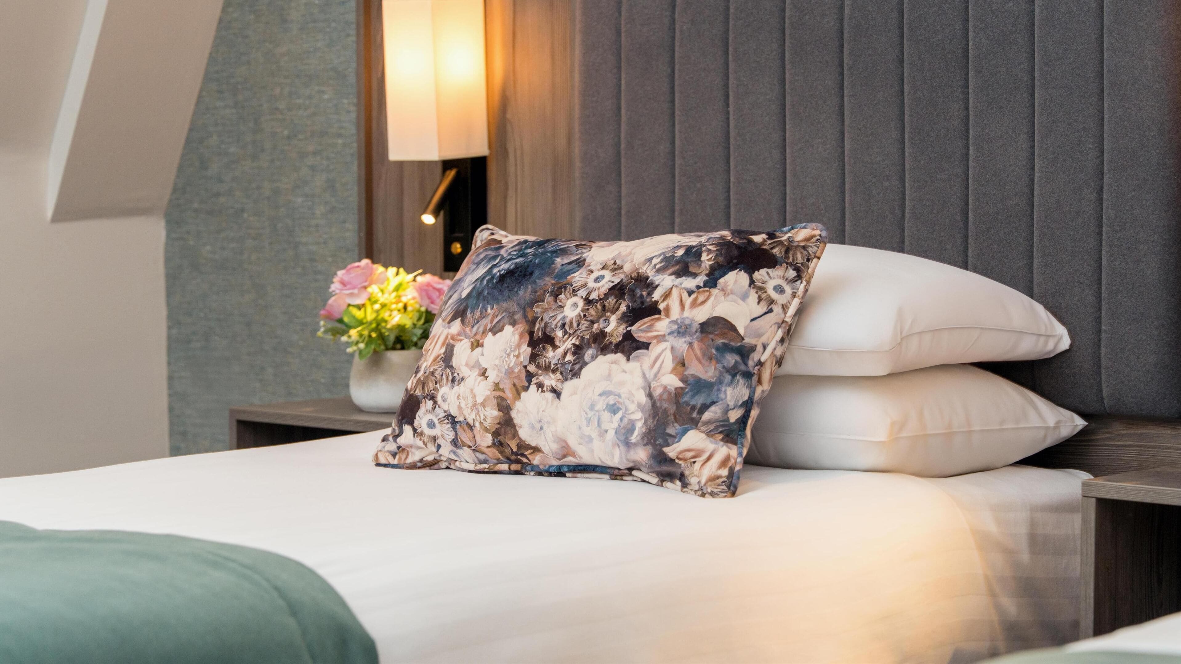 A neatly made bed with a floral pillow and a white pillow, a bedside table with a lamp, and a vase with flowers.