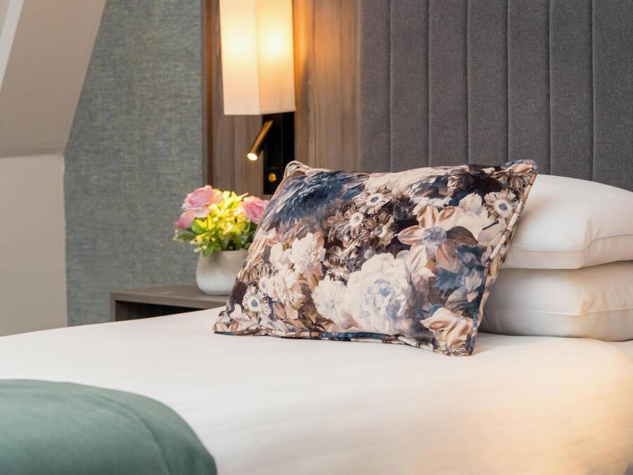 A neatly made bed with a floral pillow and a white pillow, a bedside table with a lamp, and a vase with flowers.