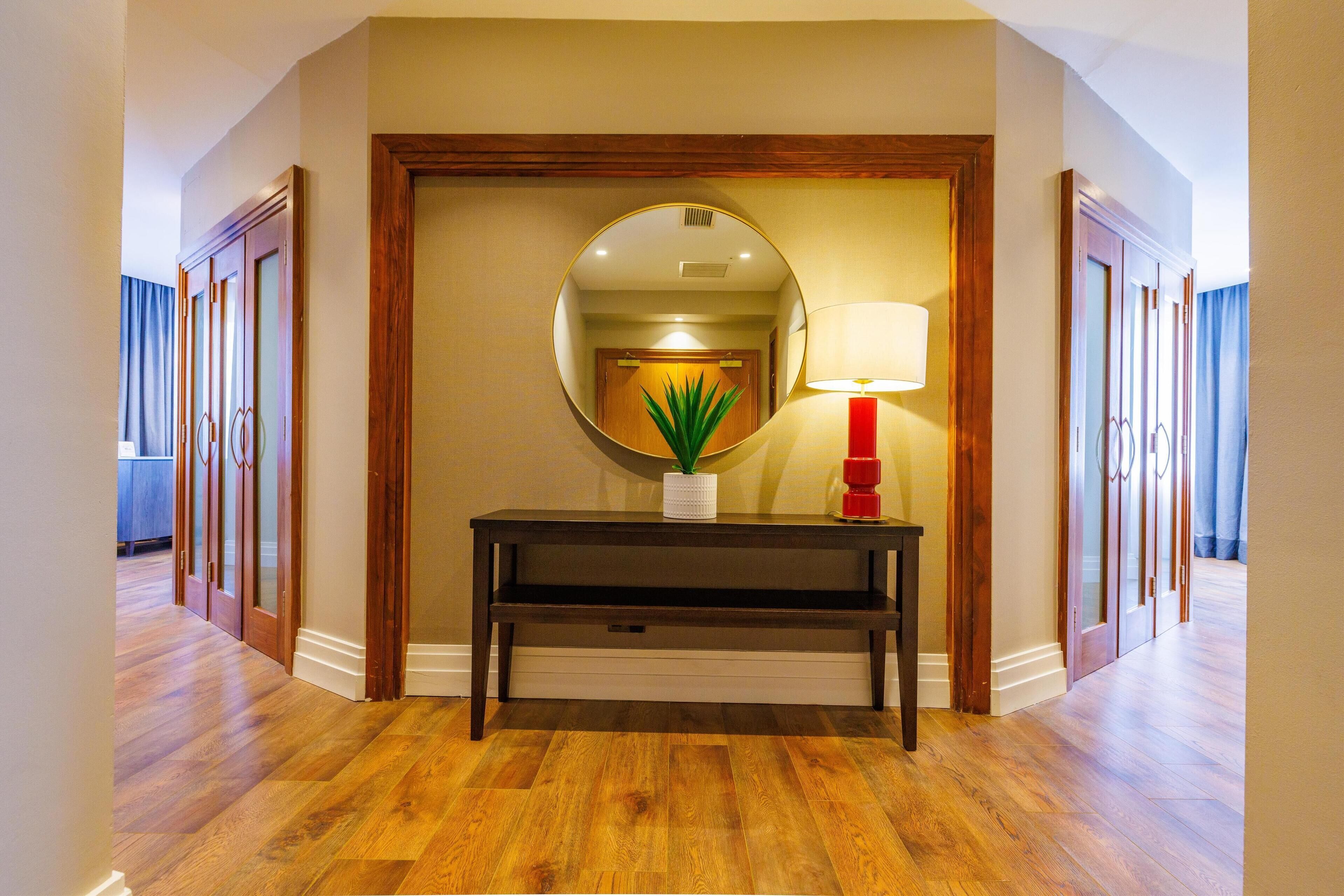 A hallway with wooden flooring, a dark console table with a potted plant, a mirror, and a red lamp.