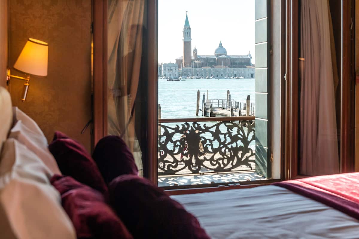 Ein Hotelzimmer mit Bett, Blick auf eine Stadtlandschaft, darunter ein markanter Turm, und einem Balkon mit Geländer.