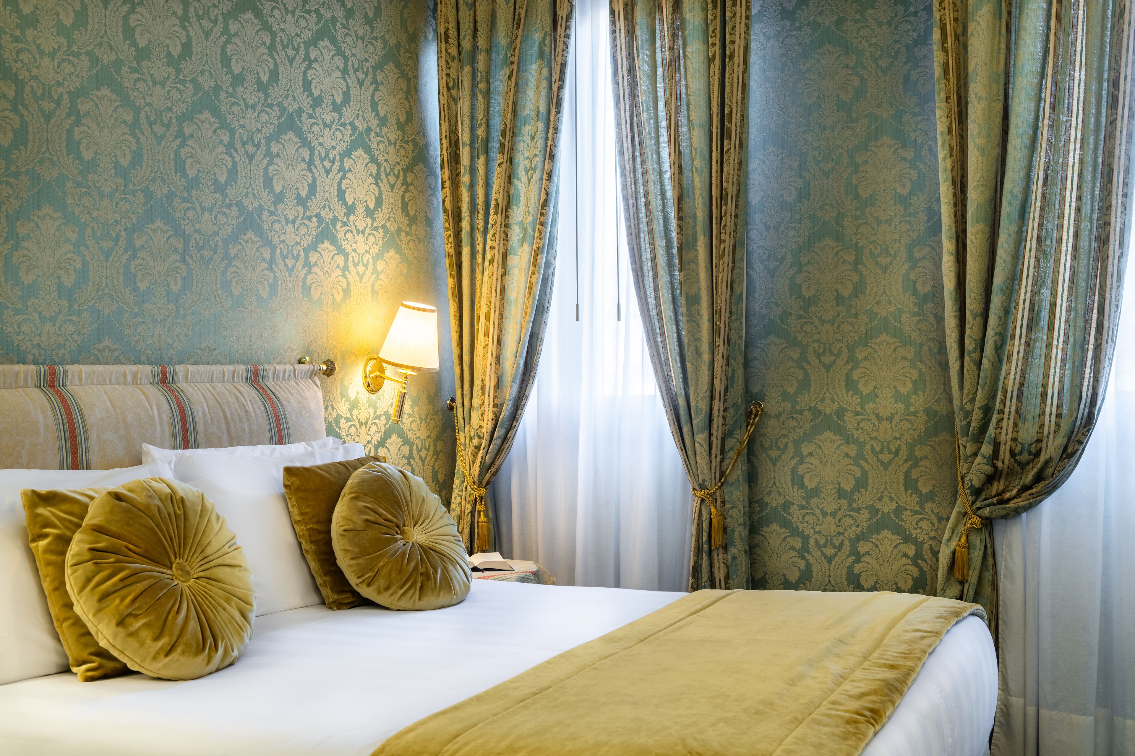 A hotel room with a bed, pillows, a nightstand, a lamp, and curtains.